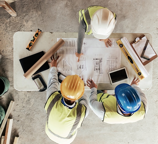 Plans de construction : l'équipe examine le projet sur le chantier. Trois professionnels du bâtiment en gilets et casques (blanc, jaune, bleu) étudient des plans d'architecture sur une table de chantier.