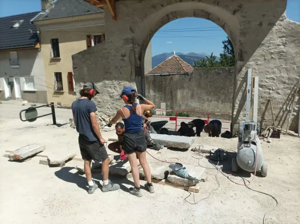 Chantier de maçonnerie : Restauration d'une arche en pierre Artisans maçons taillant la pierre devant une arche ancienne, chantier de restauration avec vue sur les montagnes.
