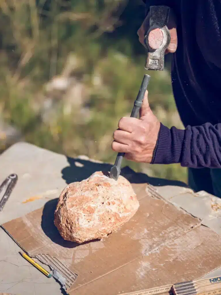 Sculpture de Pierre au Marteau et Burin : Art et Technique Un artisan sculpte une pierre poreuse avec un marteau et un burin sur un plan de travail extérieur.